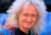Brian May smiling, with white curly hair and a colorful shirt