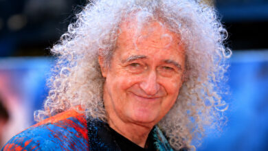 Brian May smiling, with white curly hair and a colorful shirt