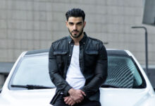 Firat Zan wearing a black jacket over a white T-shirt, seated against a white car