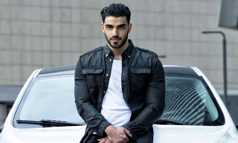 Firat Zan wearing a black jacket over a white T-shirt, seated against a white car