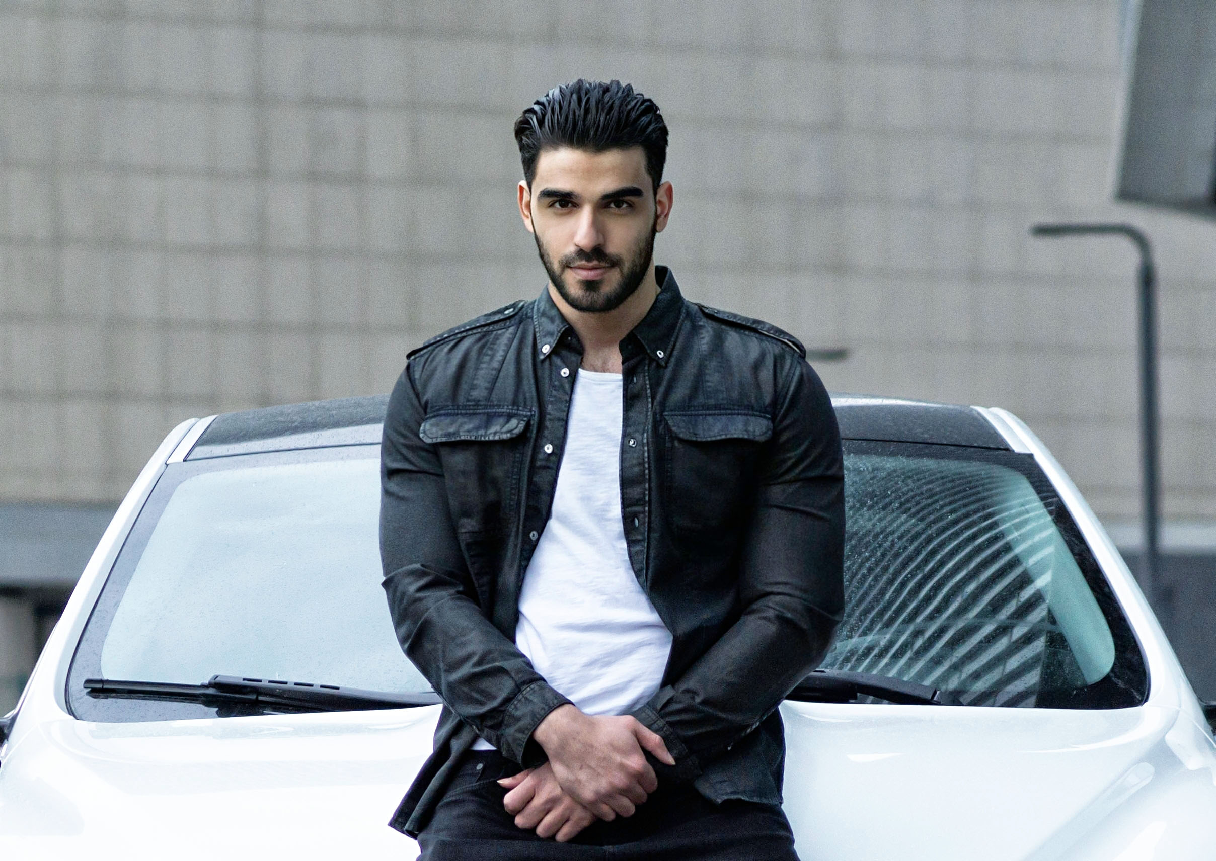 Firat Zan wearing a black jacket over a white T-shirt, seated against a white car