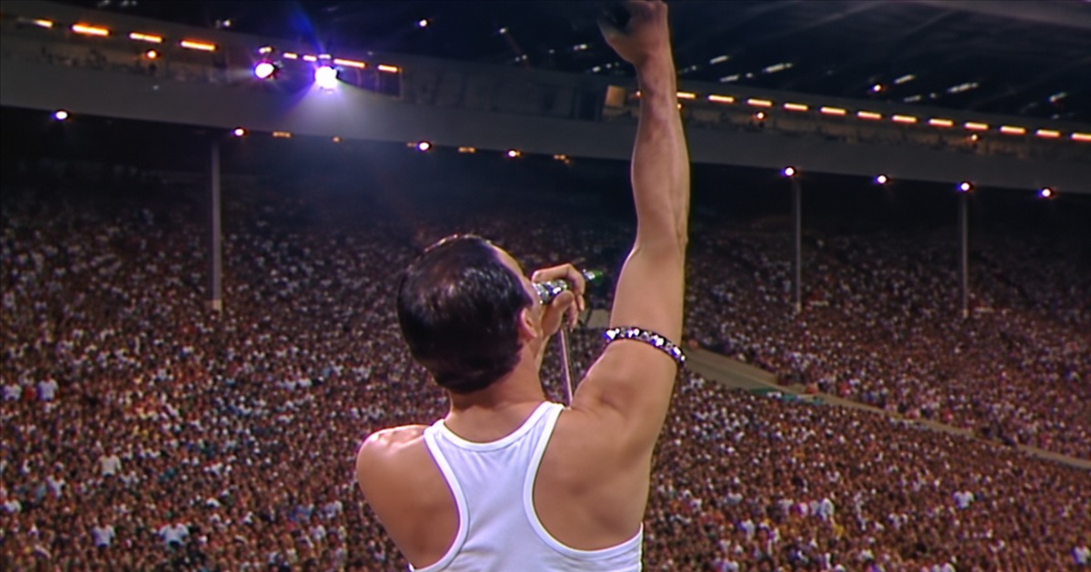 Freddie Mercury seen from behind, raising his arm towards a packed stadium.