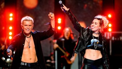 Billy Idol and Miley Cyrus raising their arms onstage under red lights at iHeartRadio.