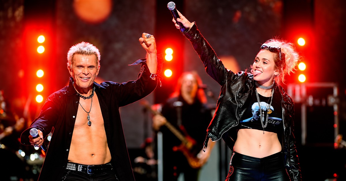 Billy Idol and Miley Cyrus raising their arms onstage under red lights at iHeartRadio.
