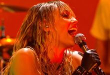 Miley Cyrus singing into a microphone under orange stage lights at the iHeartRadio Music Festival