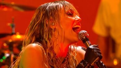 Miley Cyrus singing into a microphone under orange stage lights at the iHeartRadio Music Festival