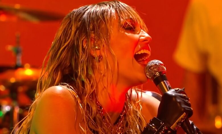 Miley Cyrus singing into a microphone under orange stage lights at the iHeartRadio Music Festival