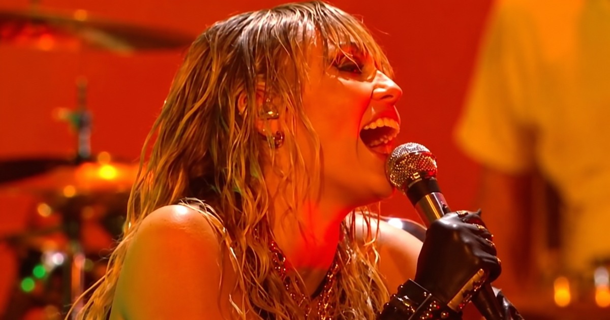 Miley Cyrus singing into a microphone under orange stage lights at the iHeartRadio Music Festival